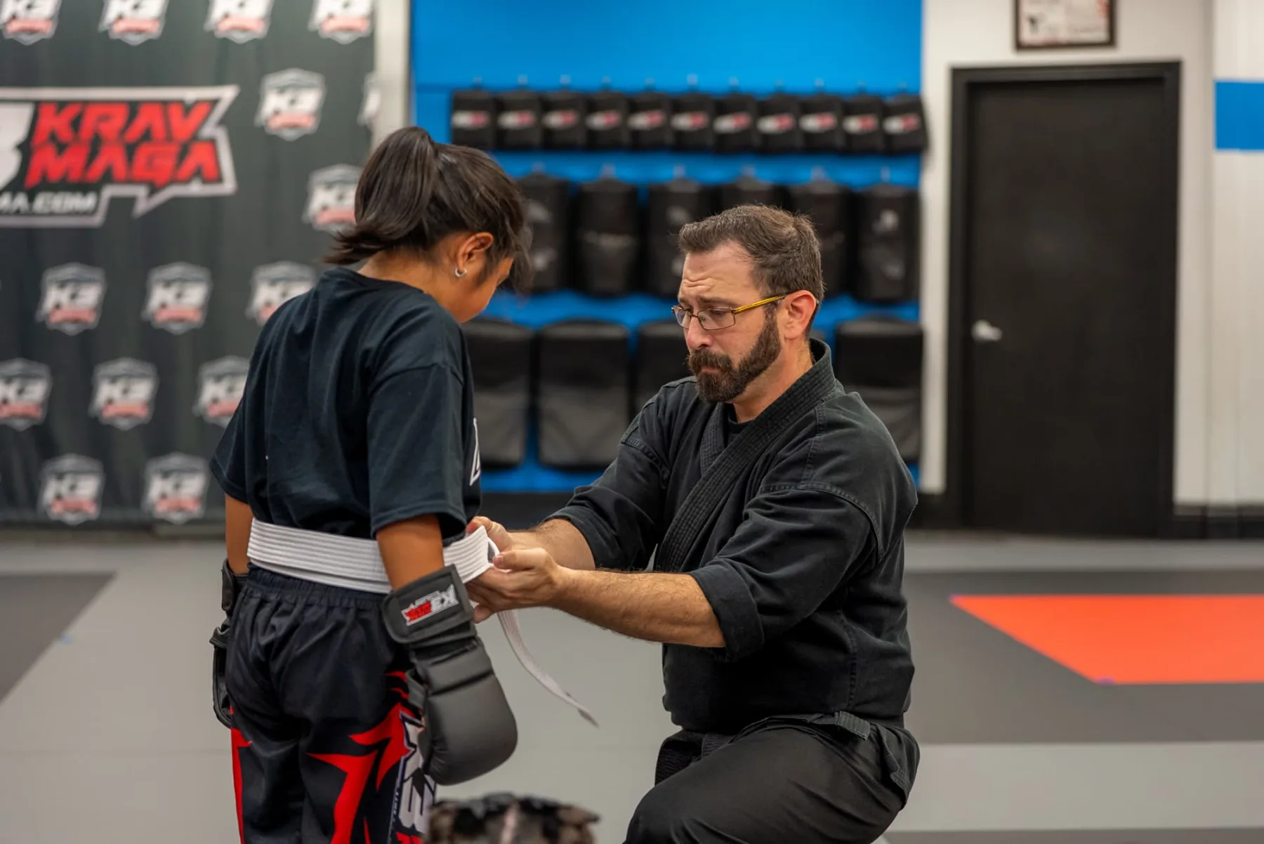 K3 instructor kneeling to tie a student's white belt